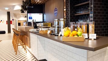 Zona de recepción de un hotel con mostrador exhibiendo alimentos para el desayuno, una pantalla de televisión y una zona de bar con estanterías.