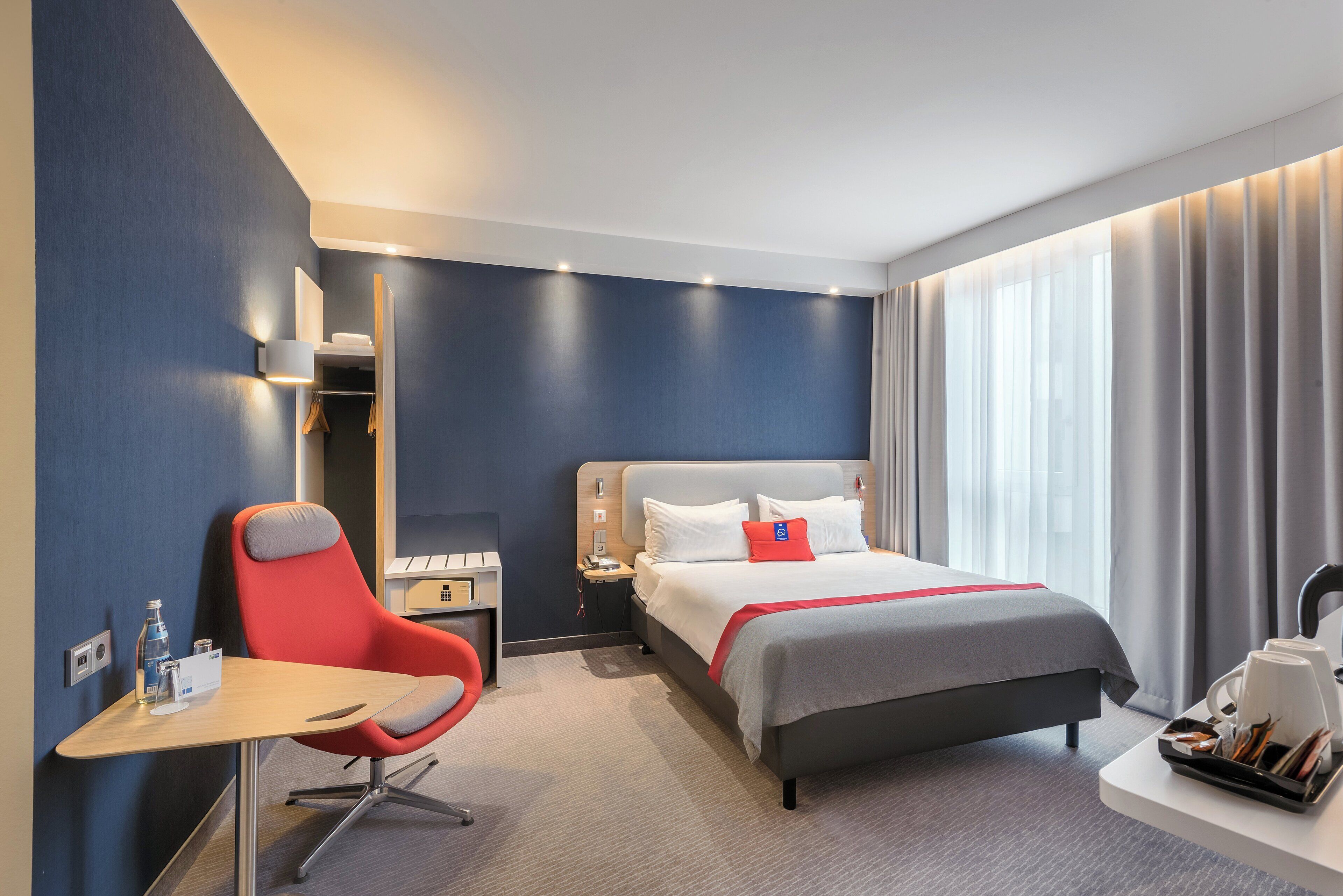 A hotel room with a bed, a desk, a chair, a nightstand, and a window with curtains.