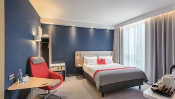 Une chambre d’hôtel comprenant un lit, un bureau, une chaise, une table de chevet et une fenêtre avec des rideaux.