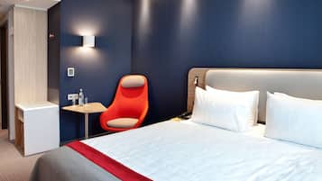 Quarto de hotel moderno com uma cama grande, uma pequena mesa, uma cadeira vermelha e uma luminária fixada na parede.