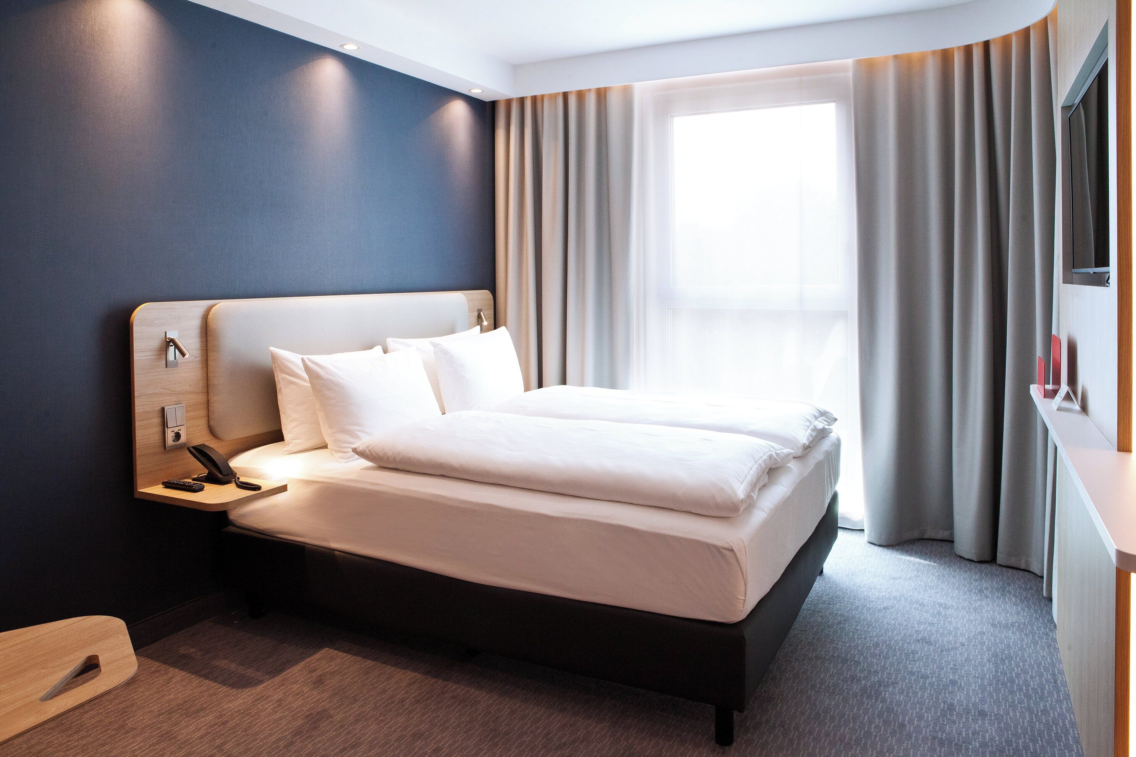 Una habitación de hotel moderna con una cama grande, una mesita de noche con teléfono y una ventana con cortinas.