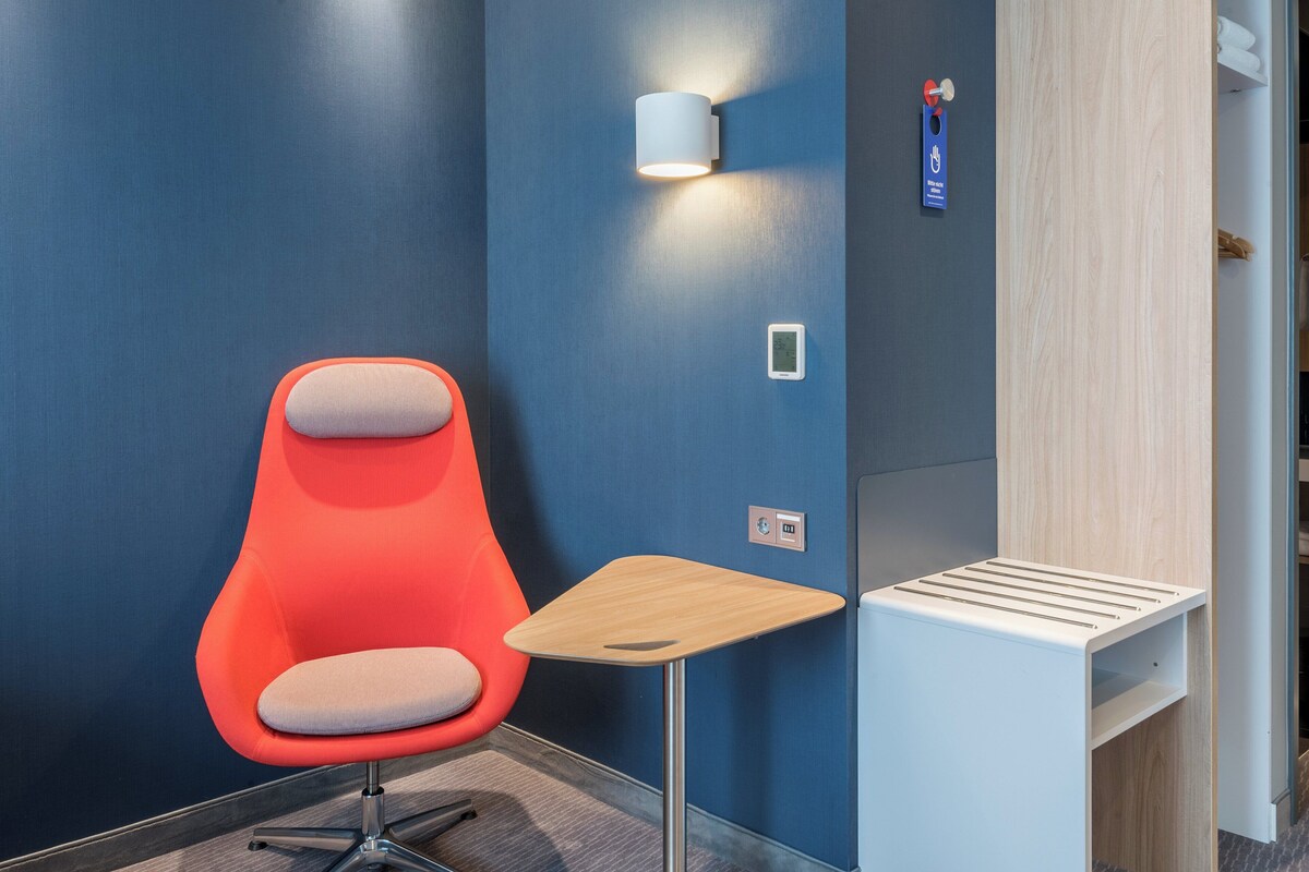 A modern room with a blue wall, a red chair, a small wooden table, a wall-mounted light, and a thermostat.