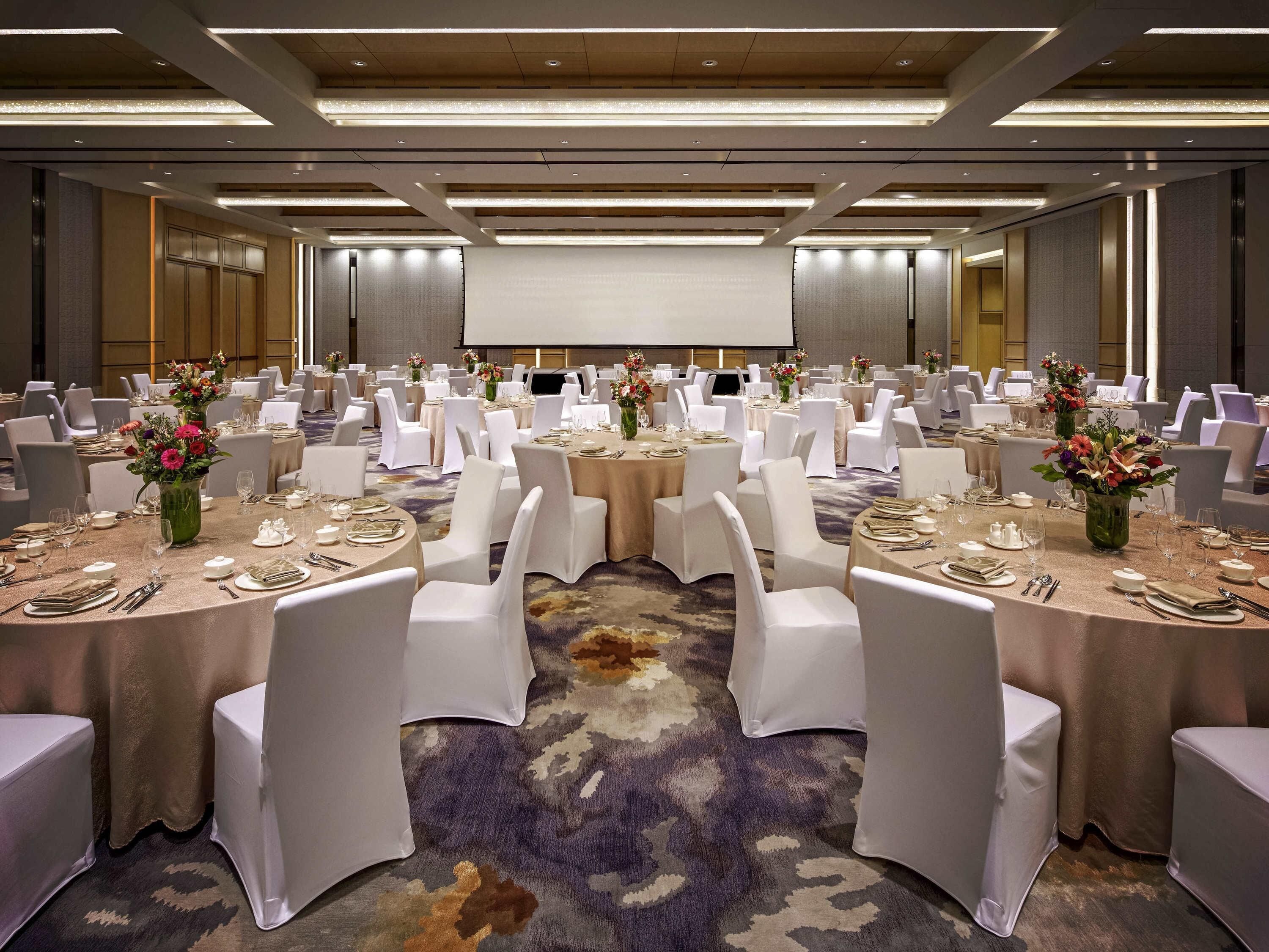 Une grande salle de banquet avec des tables rondes, des chaises blanches et des centres de table ornés de compositions florales.