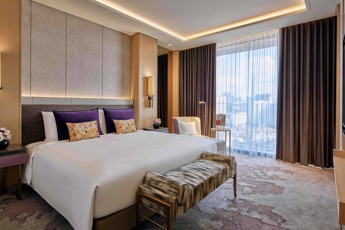 Ein modernes Hotelzimmer mit einem großen Bett, einer Bank, einem Nachttisch, einer Lampe und Blick auf die Stadt.