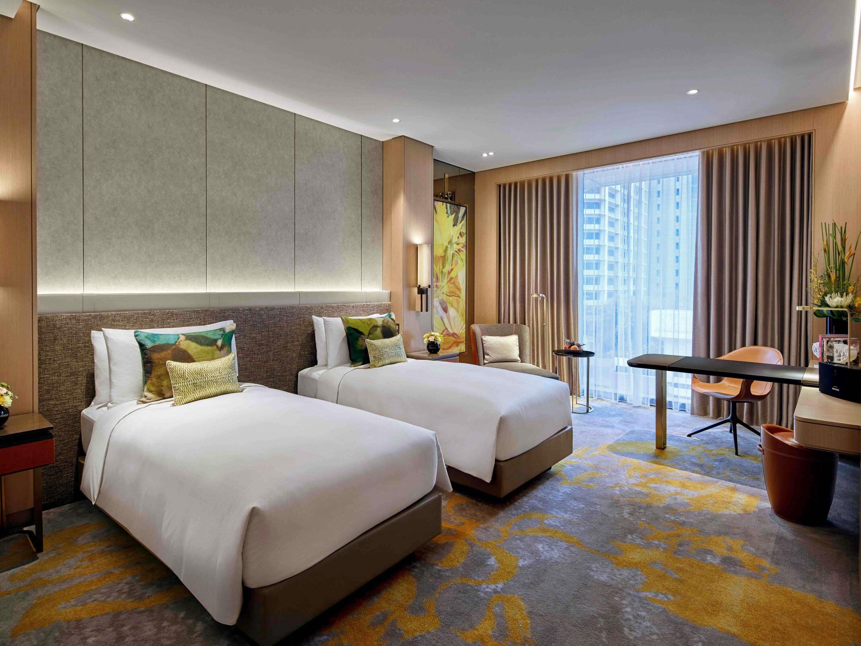 A hotel room with two beds, a desk, a chair, and a large window with curtains.