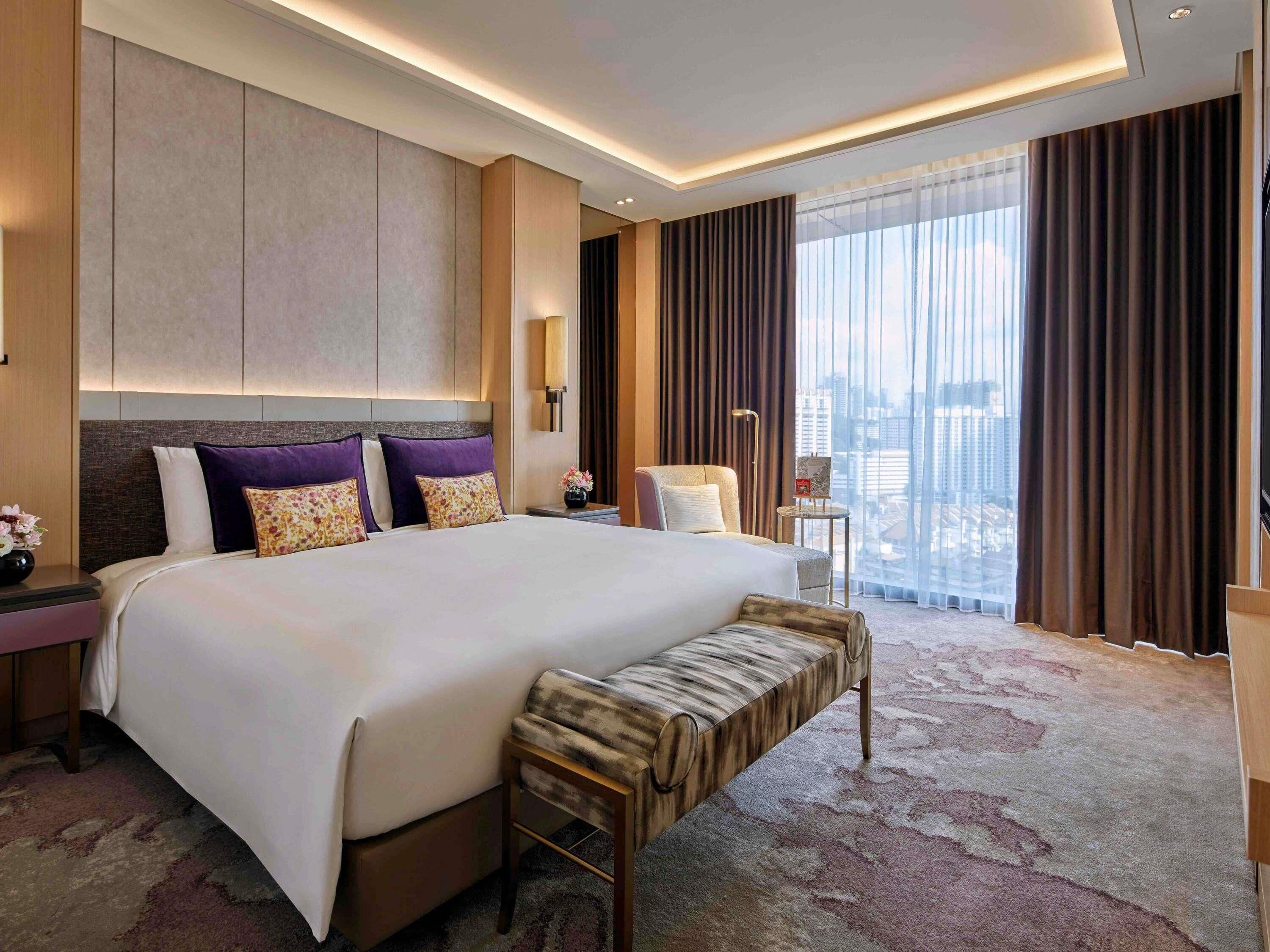 A modern hotel room with a large bed, a bench, a desk, and a view of the city.