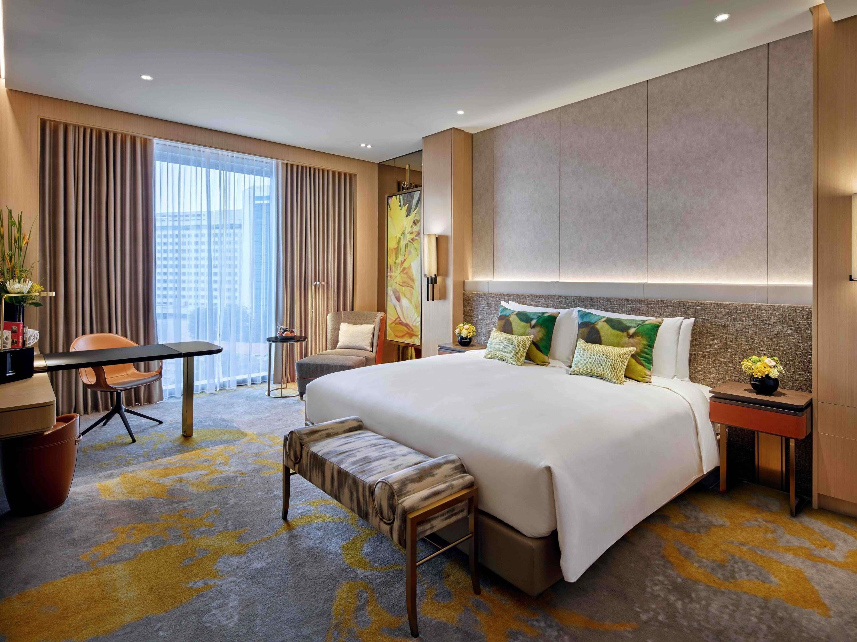 A hotel room with a large bed, a desk, a chair, a bench, and a view of the city.