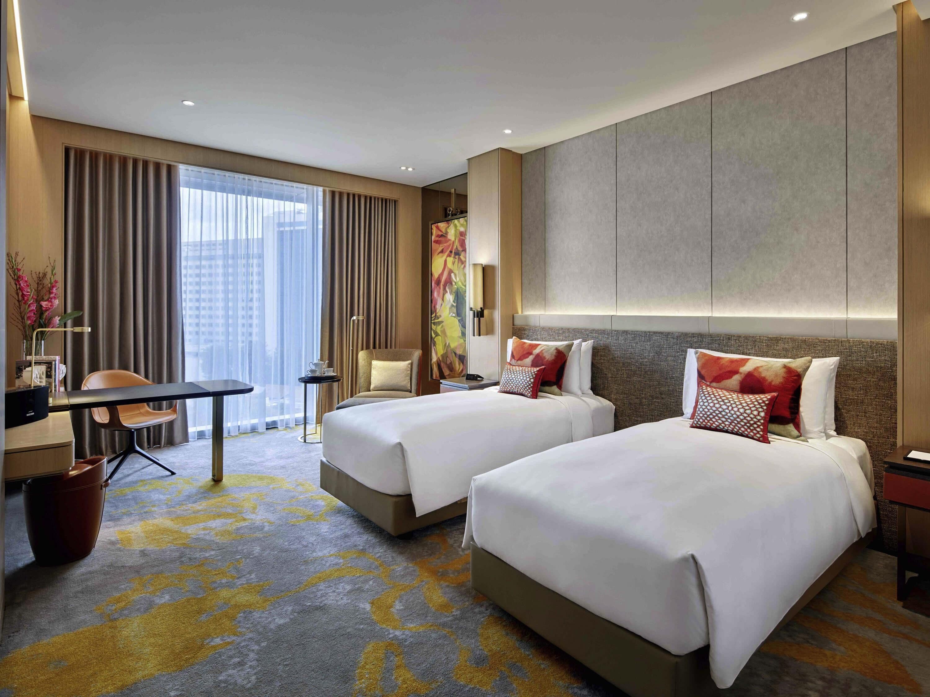 A hotel room with two beds, a desk, a chair, a small table, and a large window with curtains.
