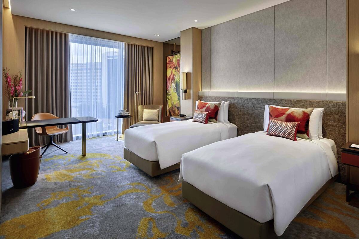 Ein Hotelzimmer mit zwei Betten, einem Schreibtisch, einem Stuhl, einem kleinen Tisch und einem großen Fenster mit Vorhängen.
