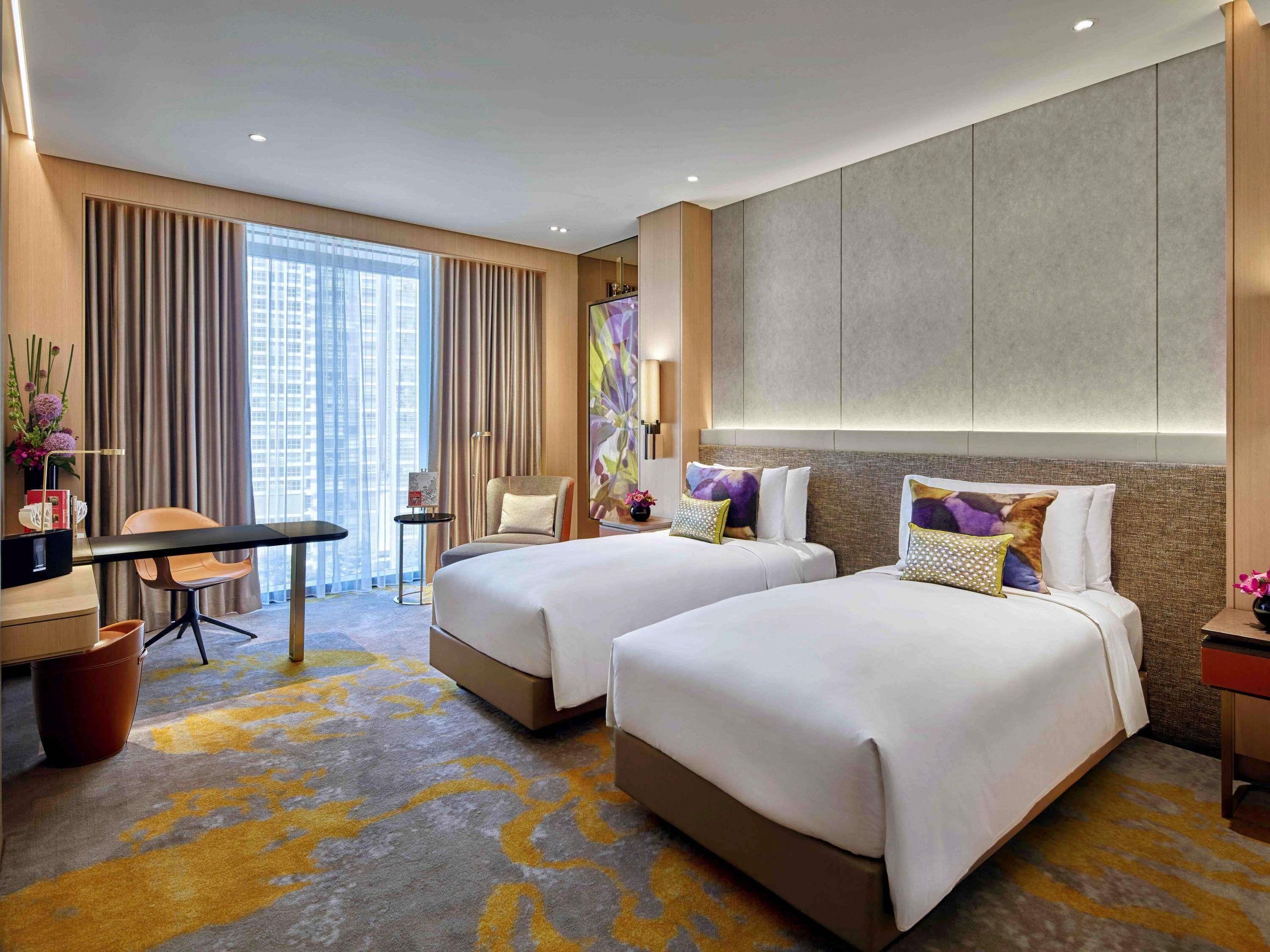 A hotel room with two beds, a desk, a chair, and a large window with curtains.