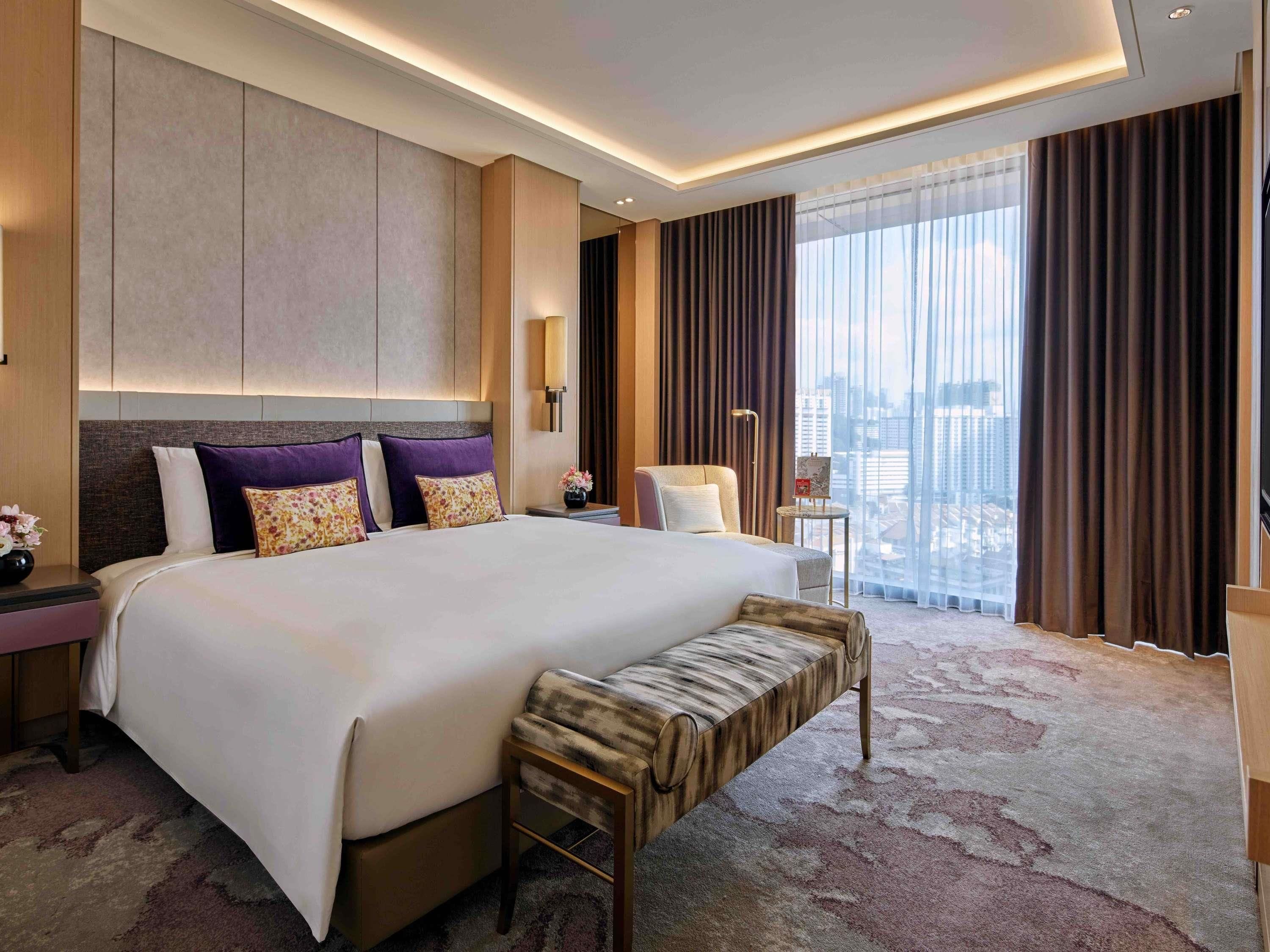 A modern hotel room with a large bed, a bench, a nightstand, a lamp, and a view of the city.