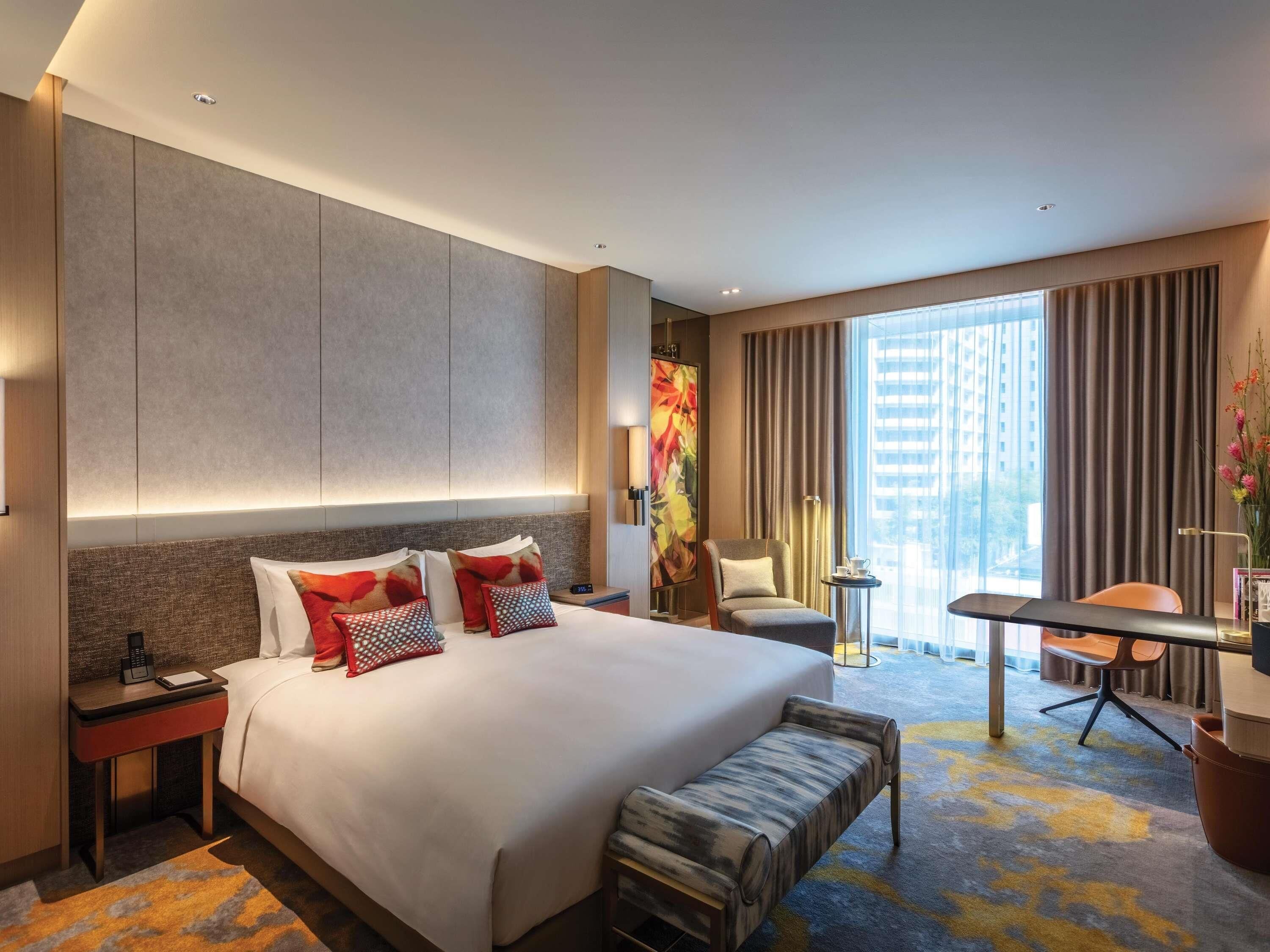 A modern hotel room with a large bed, a desk, a chair, and a view of the city.