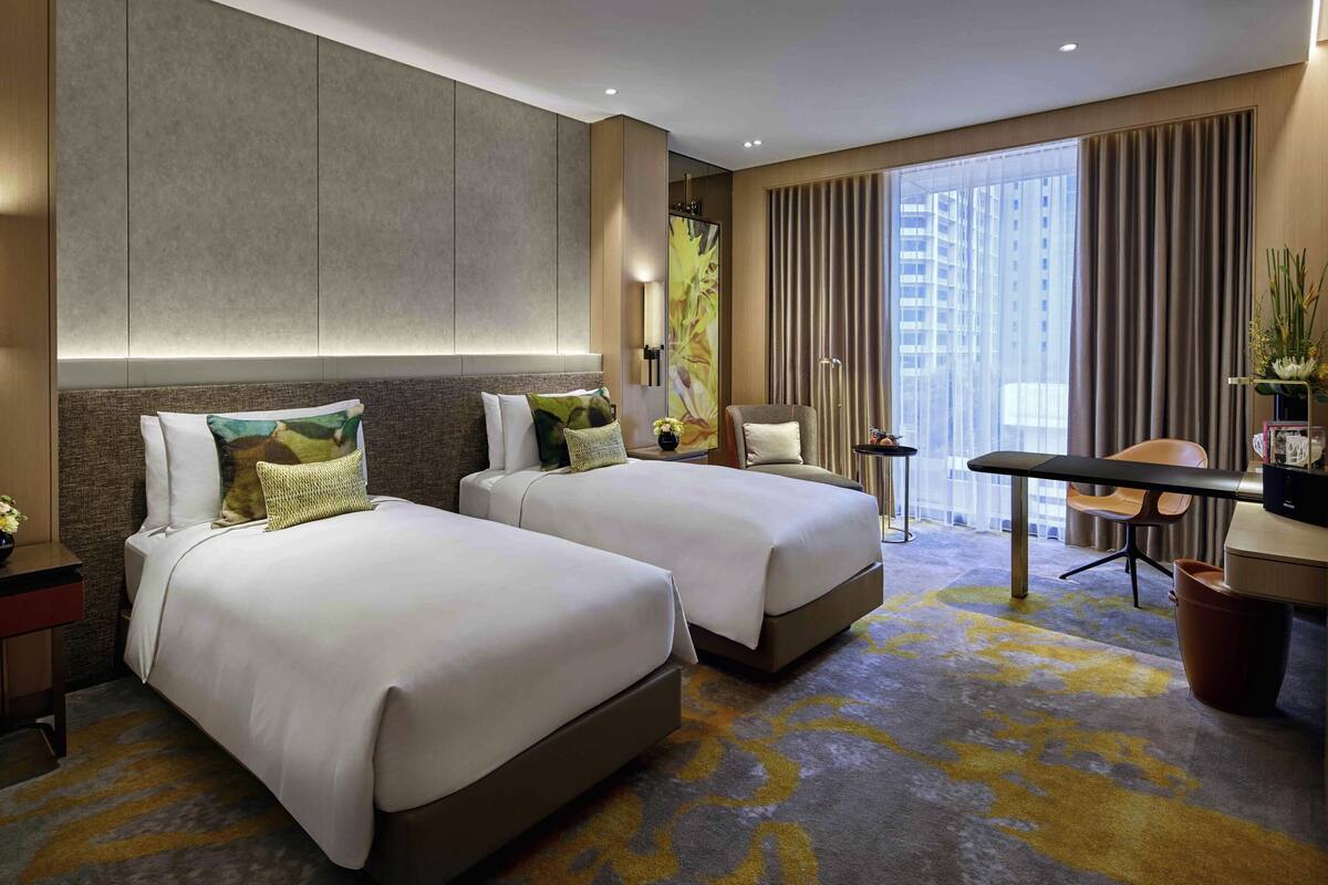 Ein Hotelzimmer mit zwei Betten, einem Schreibtisch, einem Stuhl, einem kleinen Tisch und einem großen Fenster mit Vorhängen.