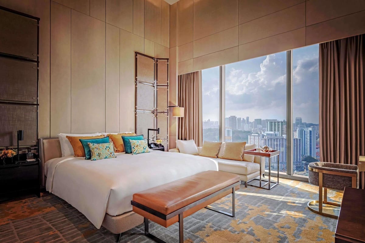 Amplia habitación de hotel con una cama grande, un banco, una pequeña mesa y vistas al horizonte de la ciudad.