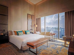 Amplia habitación de hotel con una cama grande, un banco, una pequeña mesa y vistas al horizonte de la ciudad.