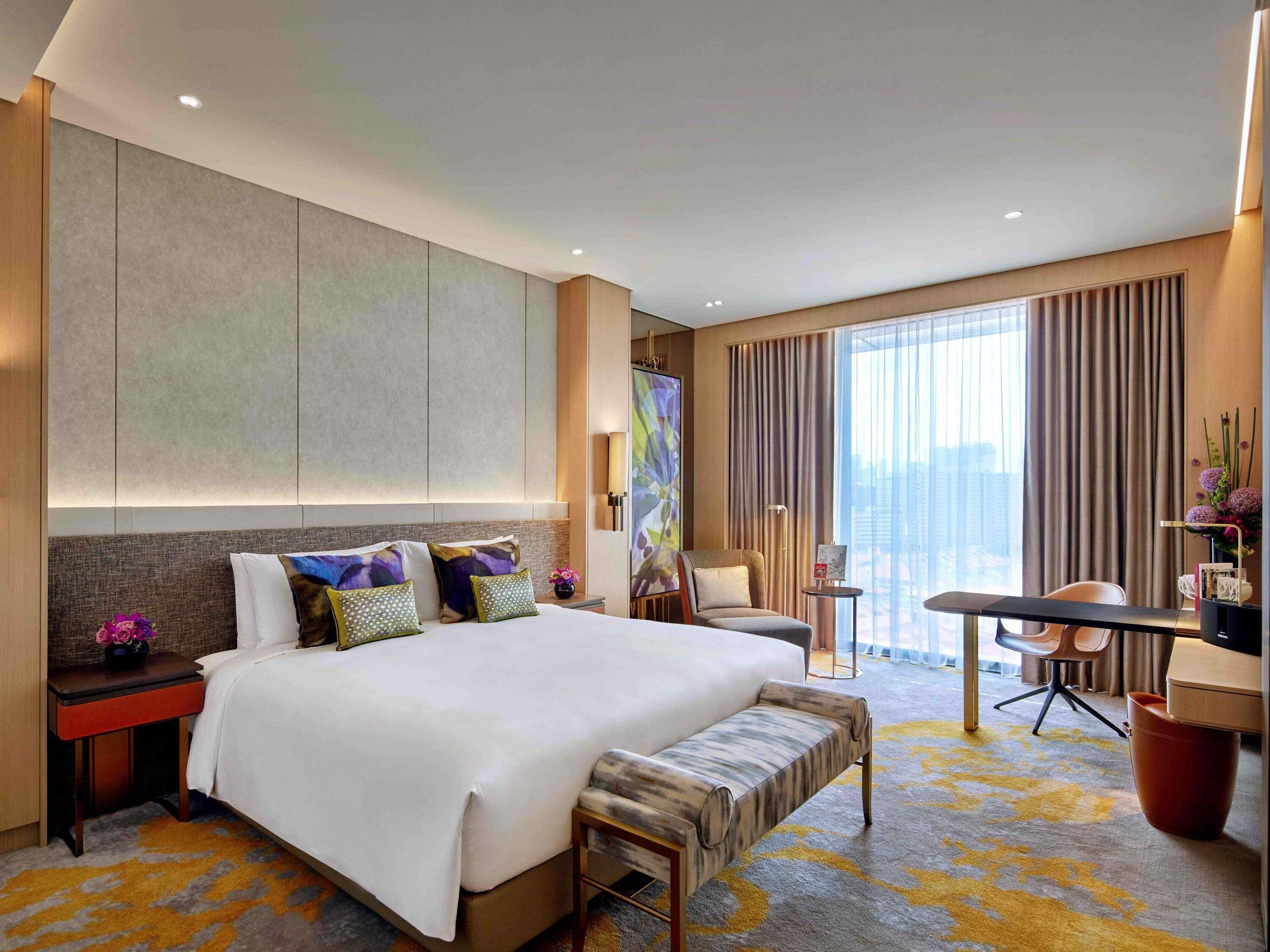 A modern hotel room with a large bed, a desk, a chair, and a view of the city.