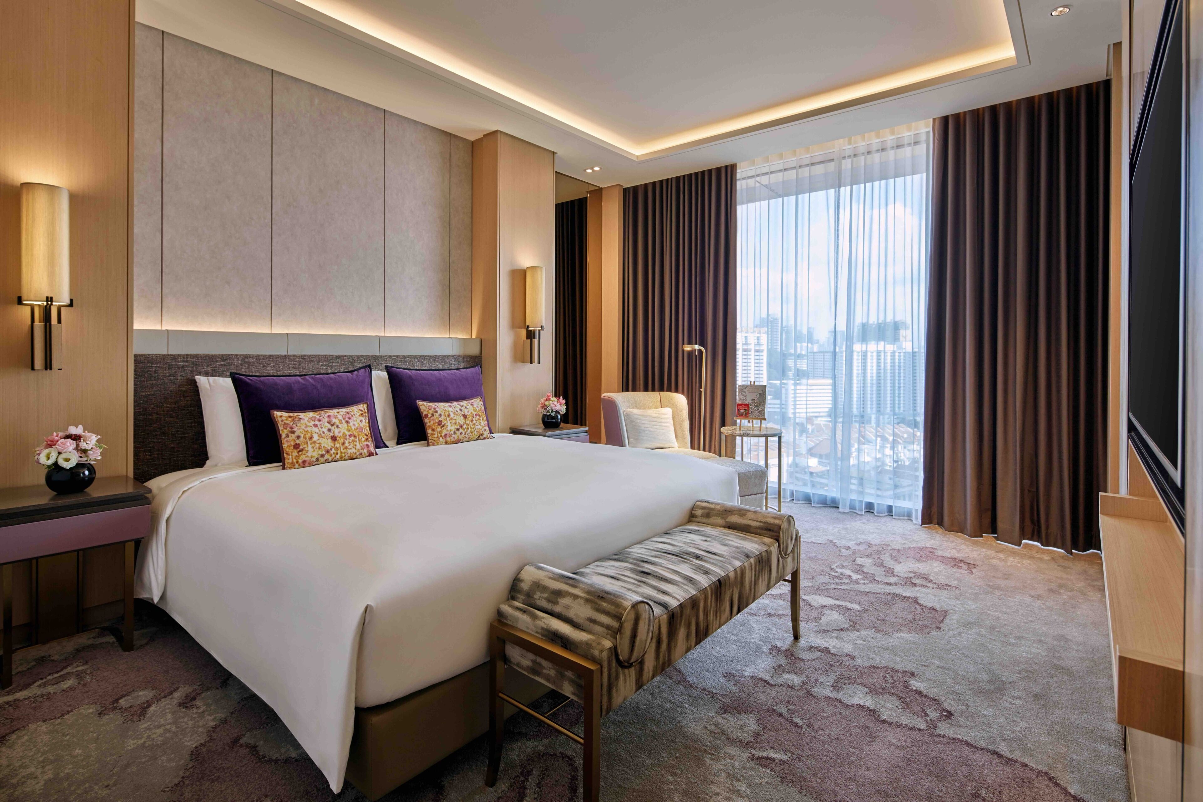 A modern hotel room with a large bed, a bench, a nightstand, a lamp, and a view of the city.