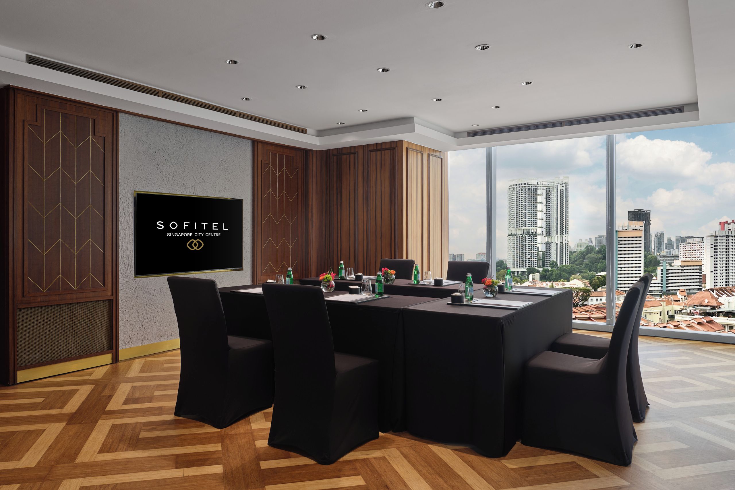 Een vergaderruimte met uitzicht op de stad, een groot scherm waarop 'SOFITEL SINGAPORE CITY CENTRE' staat, en een tafel gedekt voor vergaderingen.