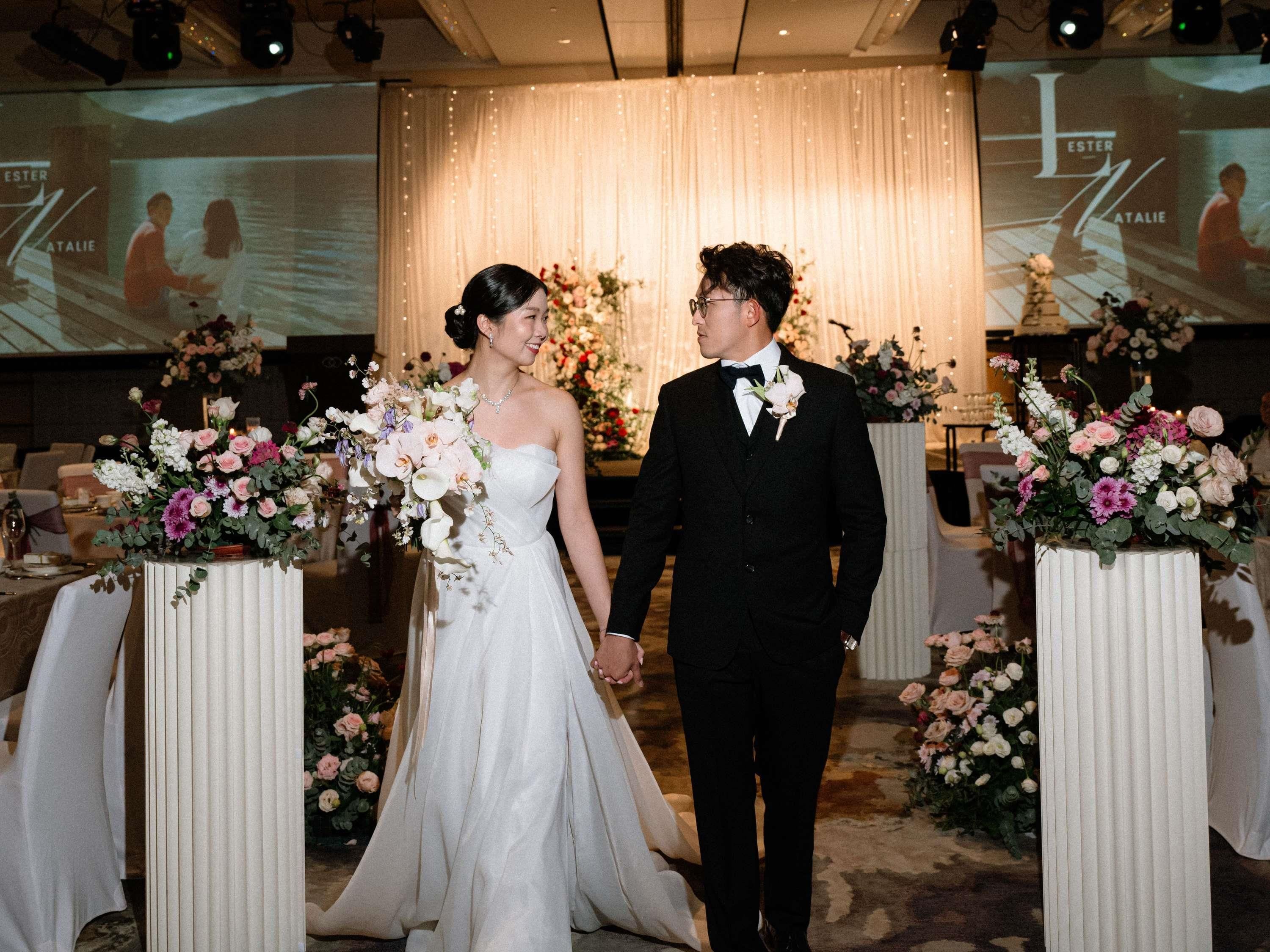 A wedding ceremony with a couple holding hands, surrounded by floral arrangements and a backdrop with a projection.