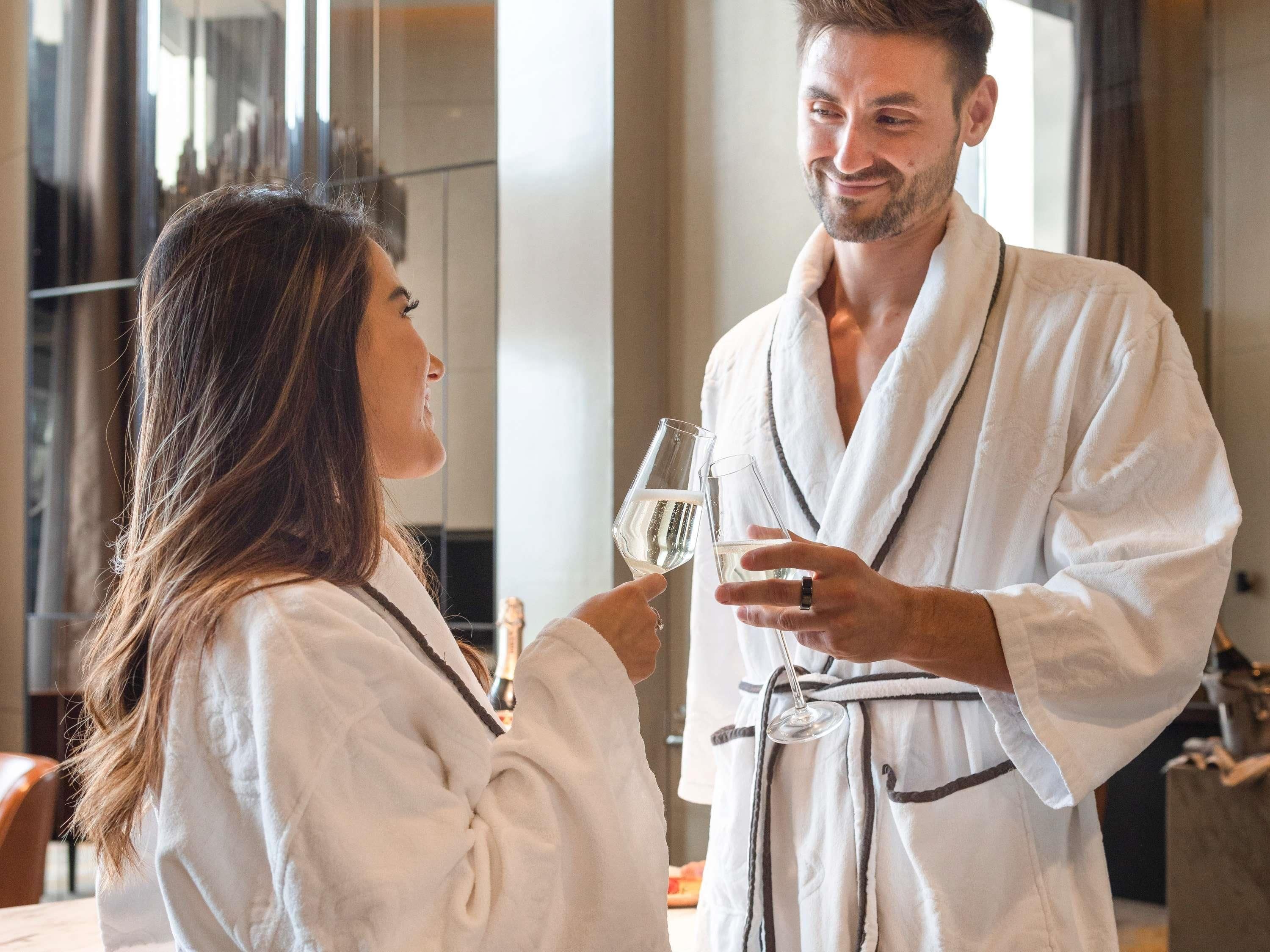 Twee personen in witte badjassen met glaasjes water, in een kamer met een modern interieur en een groot raam.