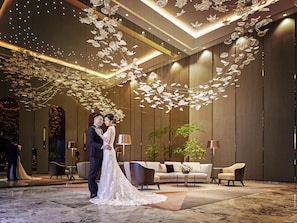 A couple in wedding attire in a modern hotel lobby with a large chandelier, seating area, and a large plant.