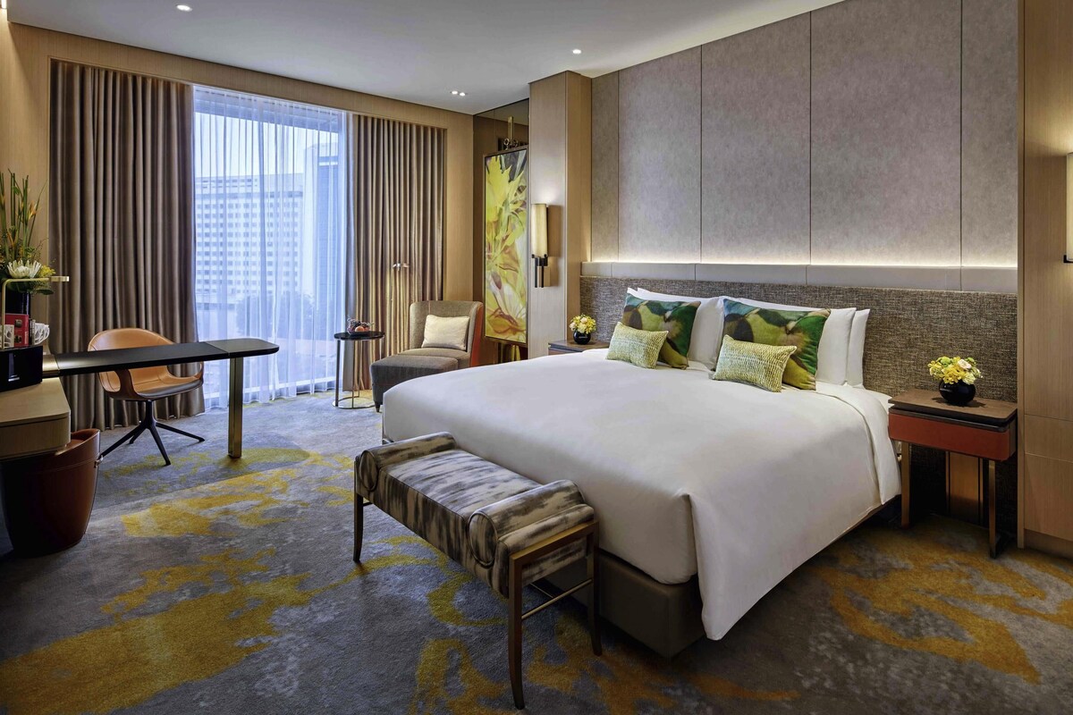 A hotel room with a large bed, a desk, a chair, a bench, and a view of the city.