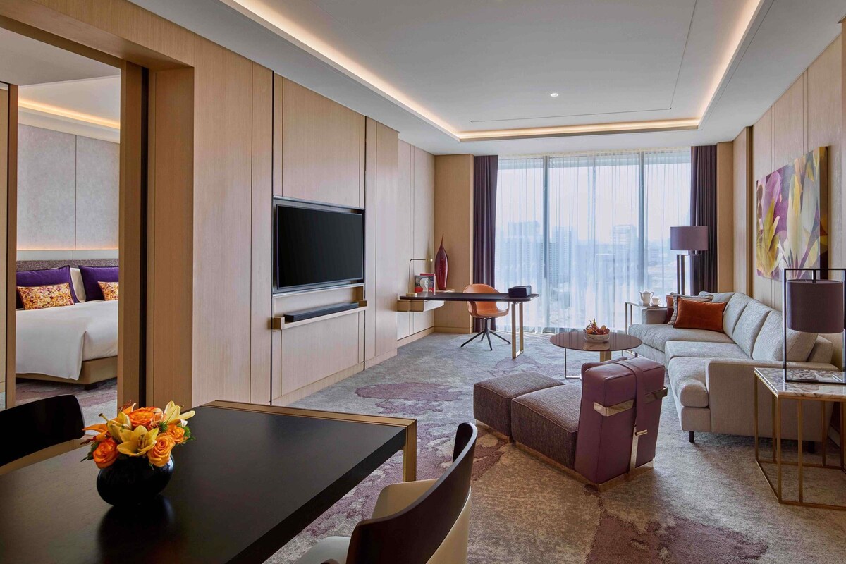 A modern hotel room with a flat-screen TV, a dining area with a table and chairs, a sofa, and a large window with a city view.