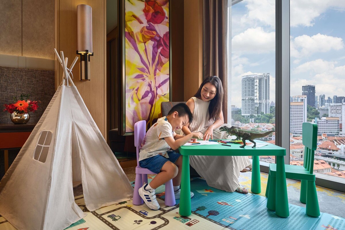 A room with a large window, a green table, a white tent, and a colorful floral painting.