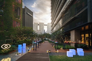 Un chemin bordé de tables et de chaises, qui mène à un bâtiment au design moderne, avec en arrière-plan la vue de gratte-ciel.