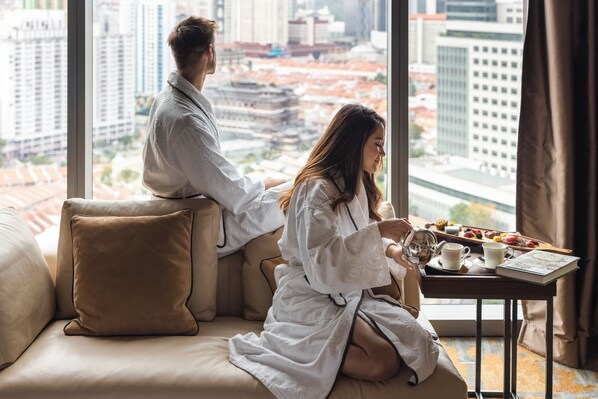 A couple in a hotel room with a city view, sitting on a sofa with a tray of pastries and a book.