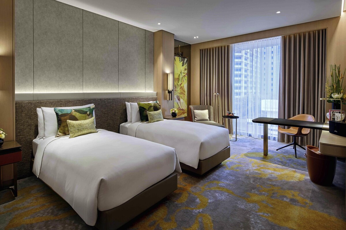 A hotel room with two beds, a desk, a chair, a small table, and a large window with curtains.