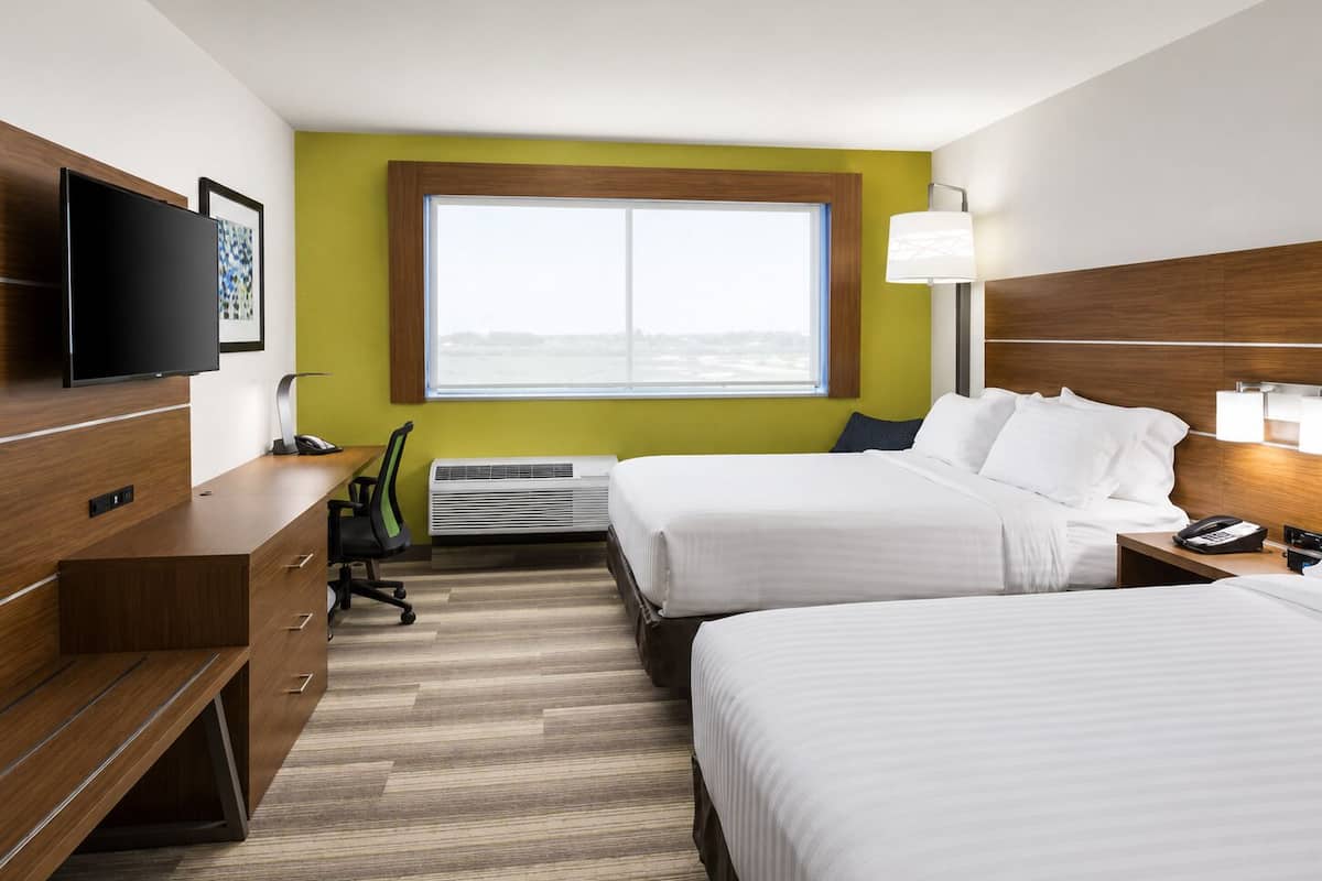 A hotel room with two beds, a desk, a chair, a TV, a window, and a lamp.