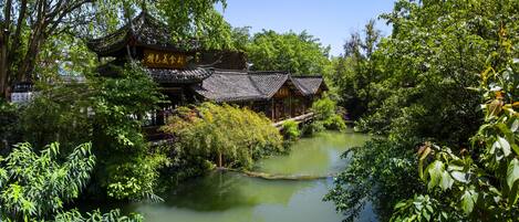 Un bâtiment traditionnel chinois, doté d’un toit recouvert de tuiles, entouré d’une végétation luxuriante et bordé d’un plan d’eau paisible.