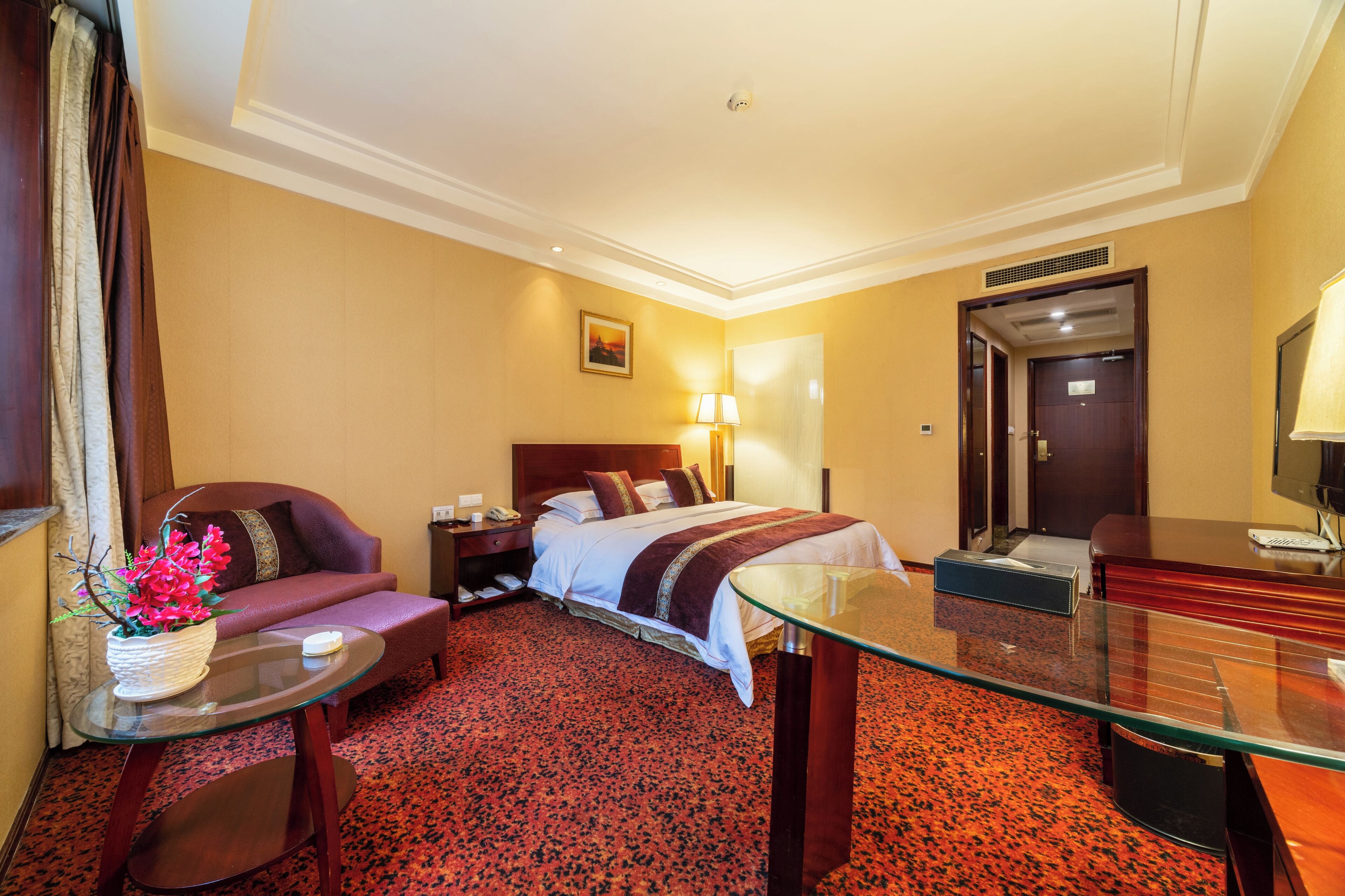 A hotel room with a bed, a sofa, a glass table, a desk, a lamp, and a painting on the wall.