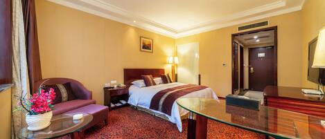A hotel room with a bed, a sofa, a glass table, a desk, a lamp, and a painting on the wall.