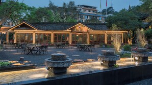 Un edificio de estilo tradicional con varias mesas y sillas dispuestas al aire libre, rodeado de un jardín con macetas de piedra y una pequeña fuente.
