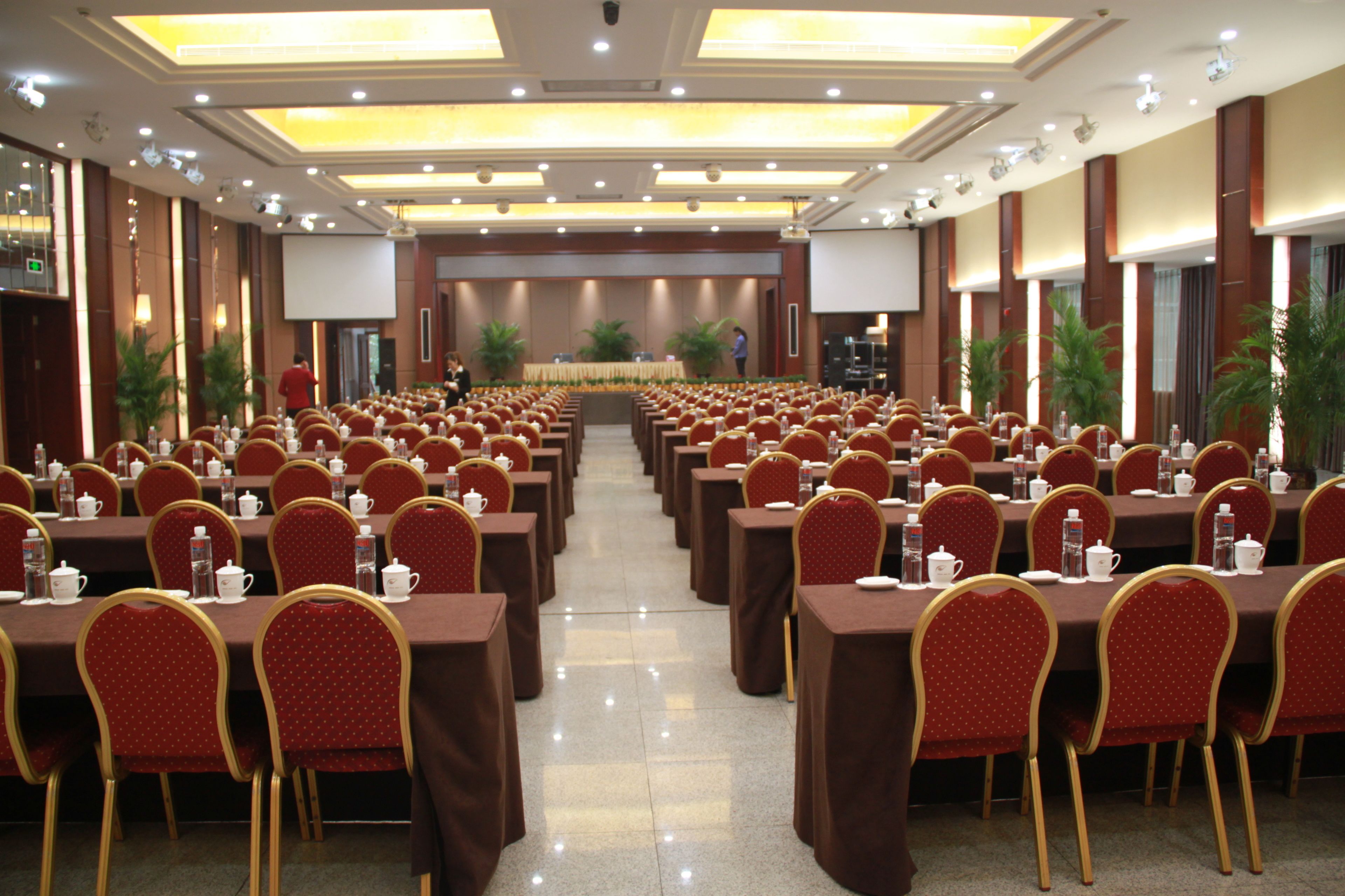 Un salón de conferencias con filas de mesas y sillas, equipado con botellas de agua y teteras, y una pantalla grande al frente.