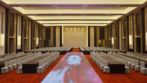 Un salón de conferencias con filas de sillas, una larga mesa y una pantalla grande que muestra una imagen floral.
