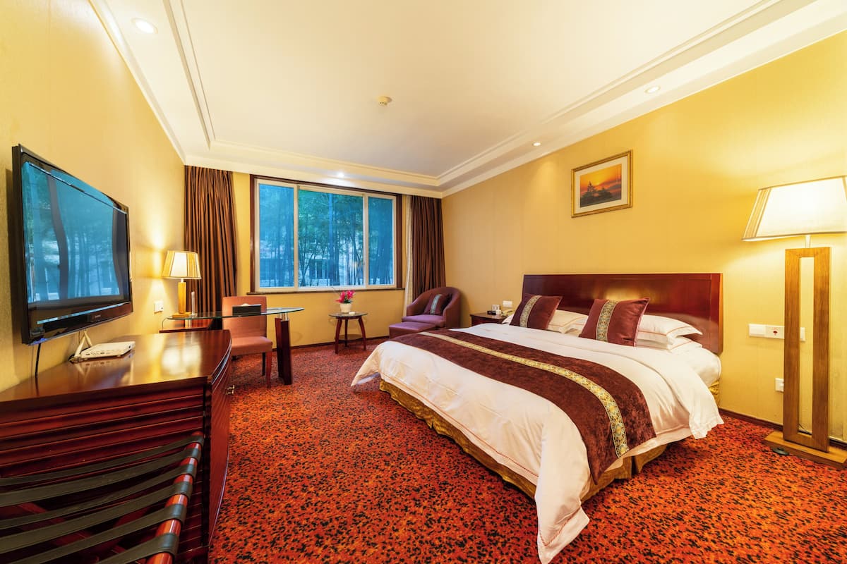 A hotel room with a large bed, a desk with a TV, a chair, a window with curtains, and a painting on the wall.