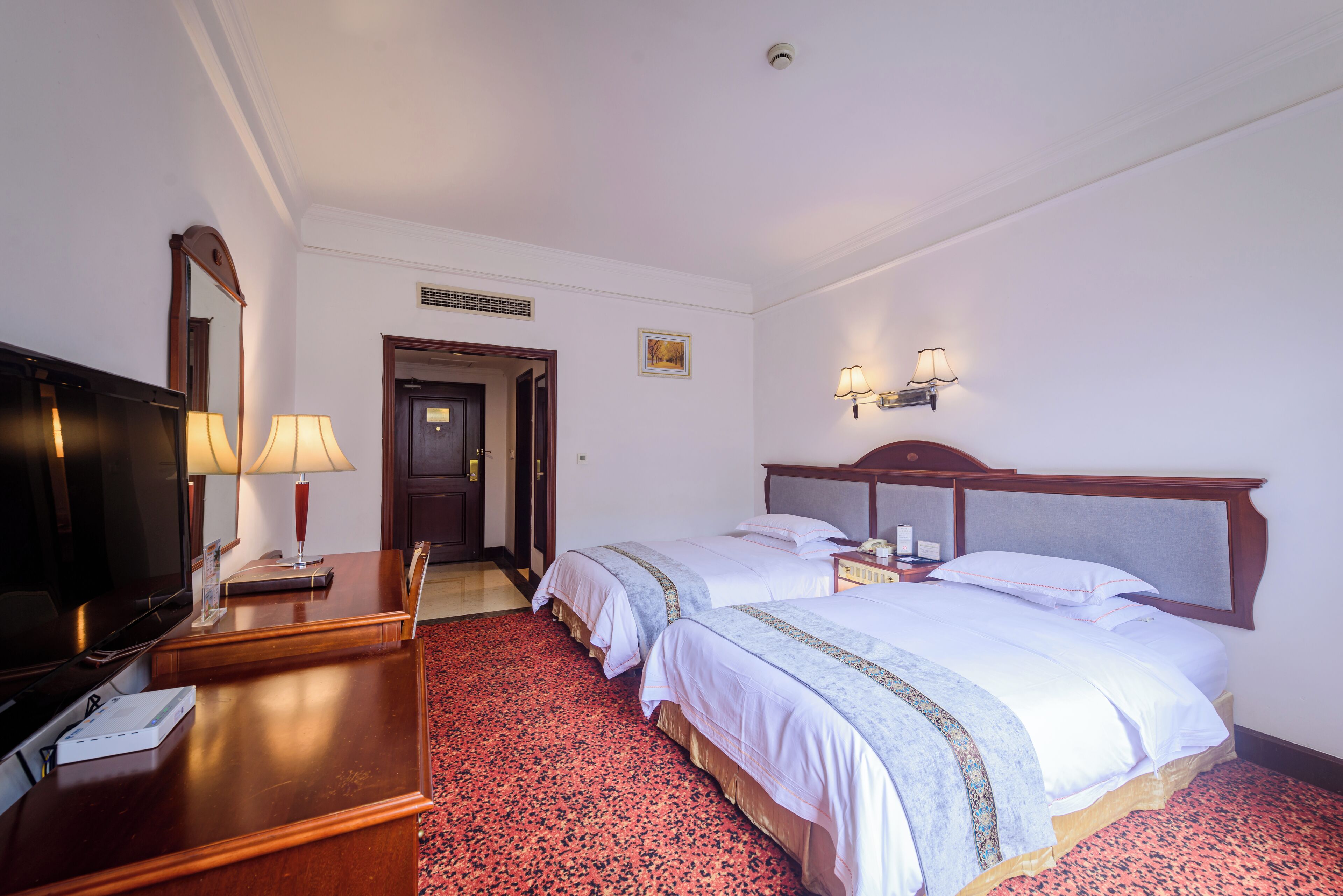 Habitación de hotel con dos camas, un escritorio y un televisor, y una alfombra roja.