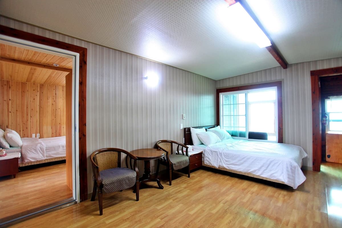 Une chambre avec un plancher en bois, un lit avec une literie blanche, deux chaises, une petite table et une fenêtre avec des stores.
