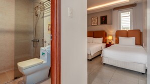 A hotel room with a shower, toilet, two beds, and a nightstand with a lamp.