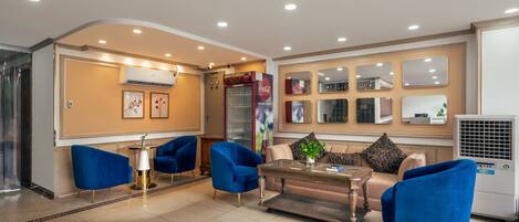 A modern lobby with blue armchairs, a coffee table, and a sofa. There are air conditioning units and framed pictures on the walls.