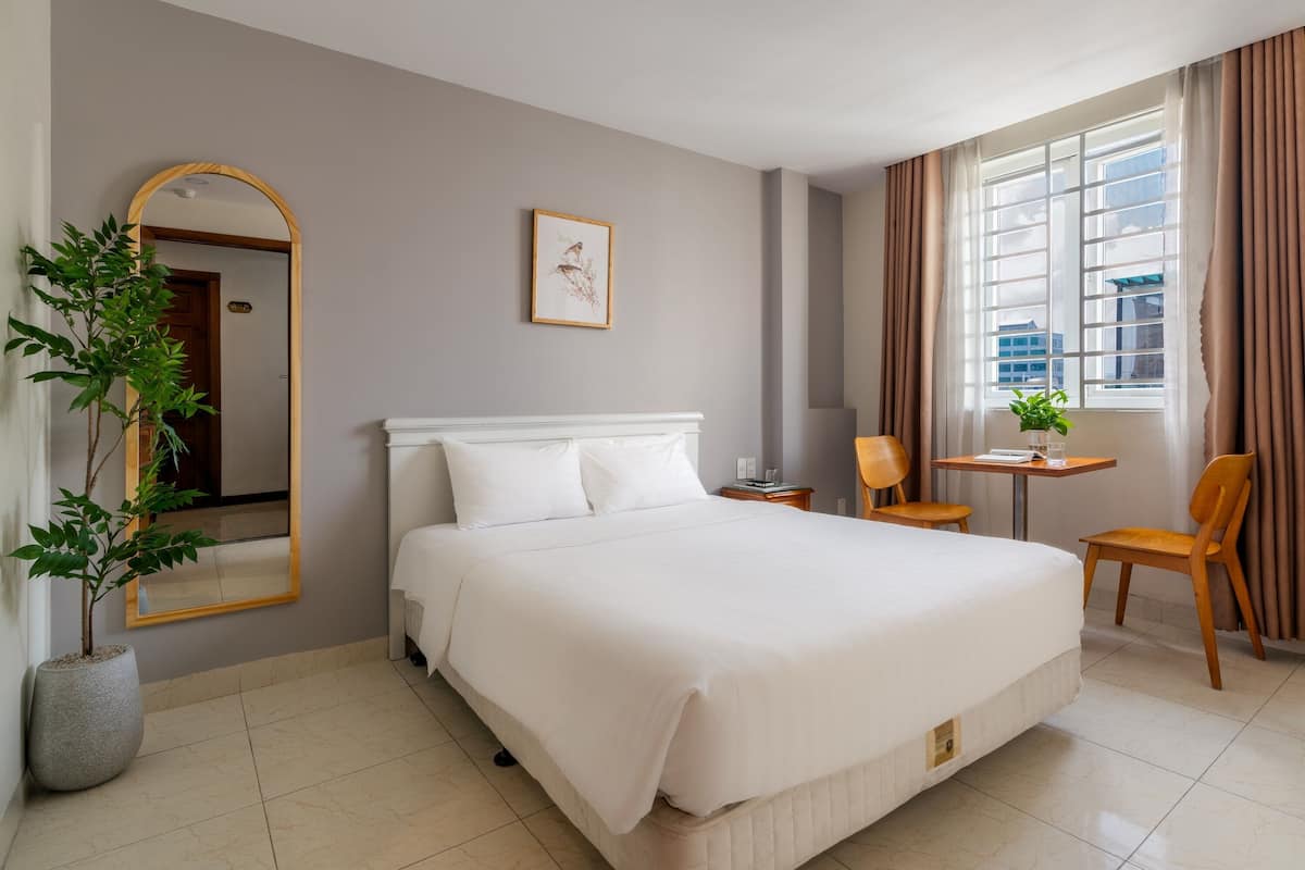 A hotel room with a large bed, a small table with a chair, a potted plant, a framed picture on the wall, and a window with curtains.