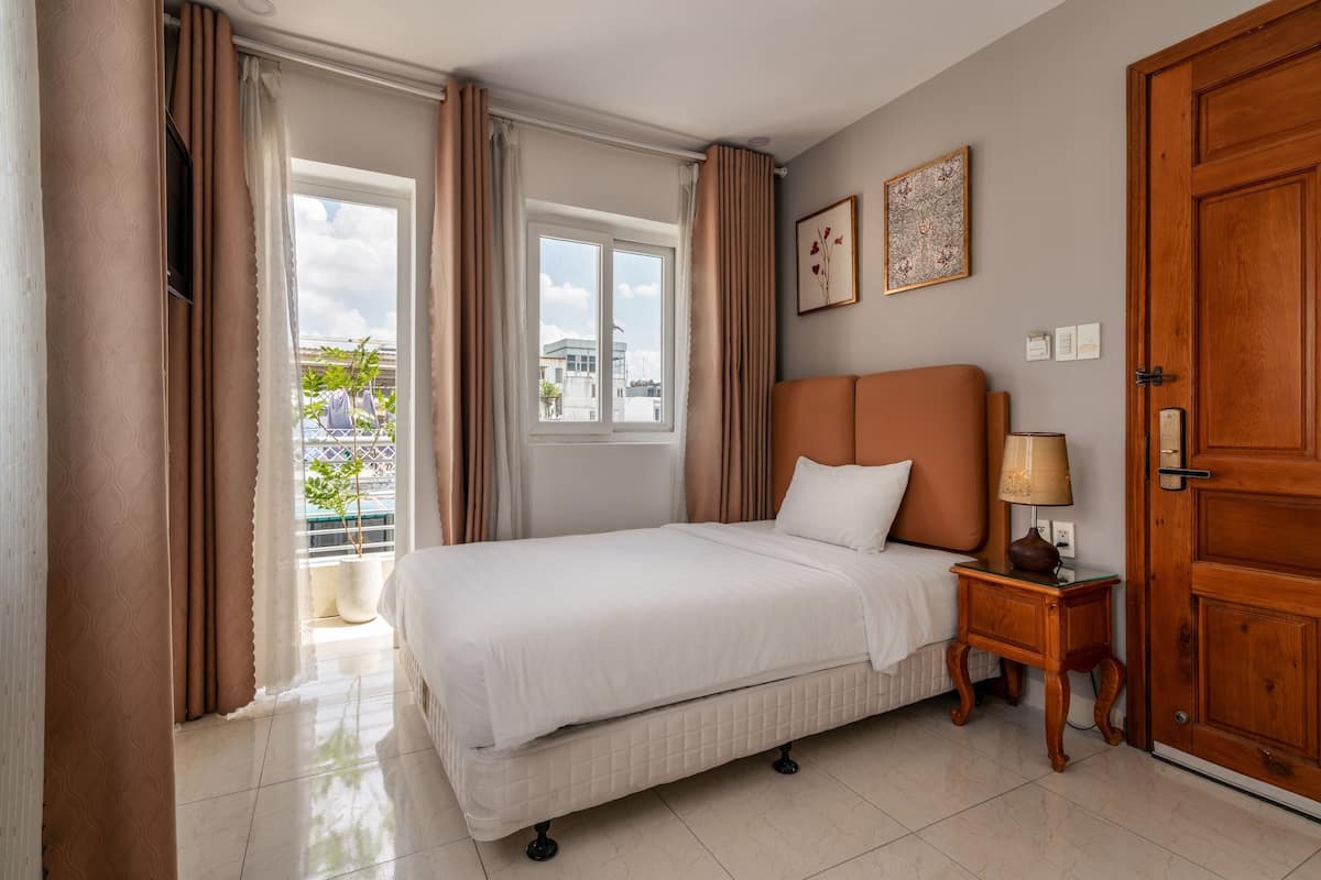 A hotel room with a bed, a nightstand, a wooden door, a window with curtains, and a balcony view.