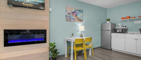 A room with a TV displaying 'Latitude 26° Waterfront Resorts,' a fireplace, a dining table with yellow chairs, a small plant, and a kitchen area with a refrigerator and microwave.