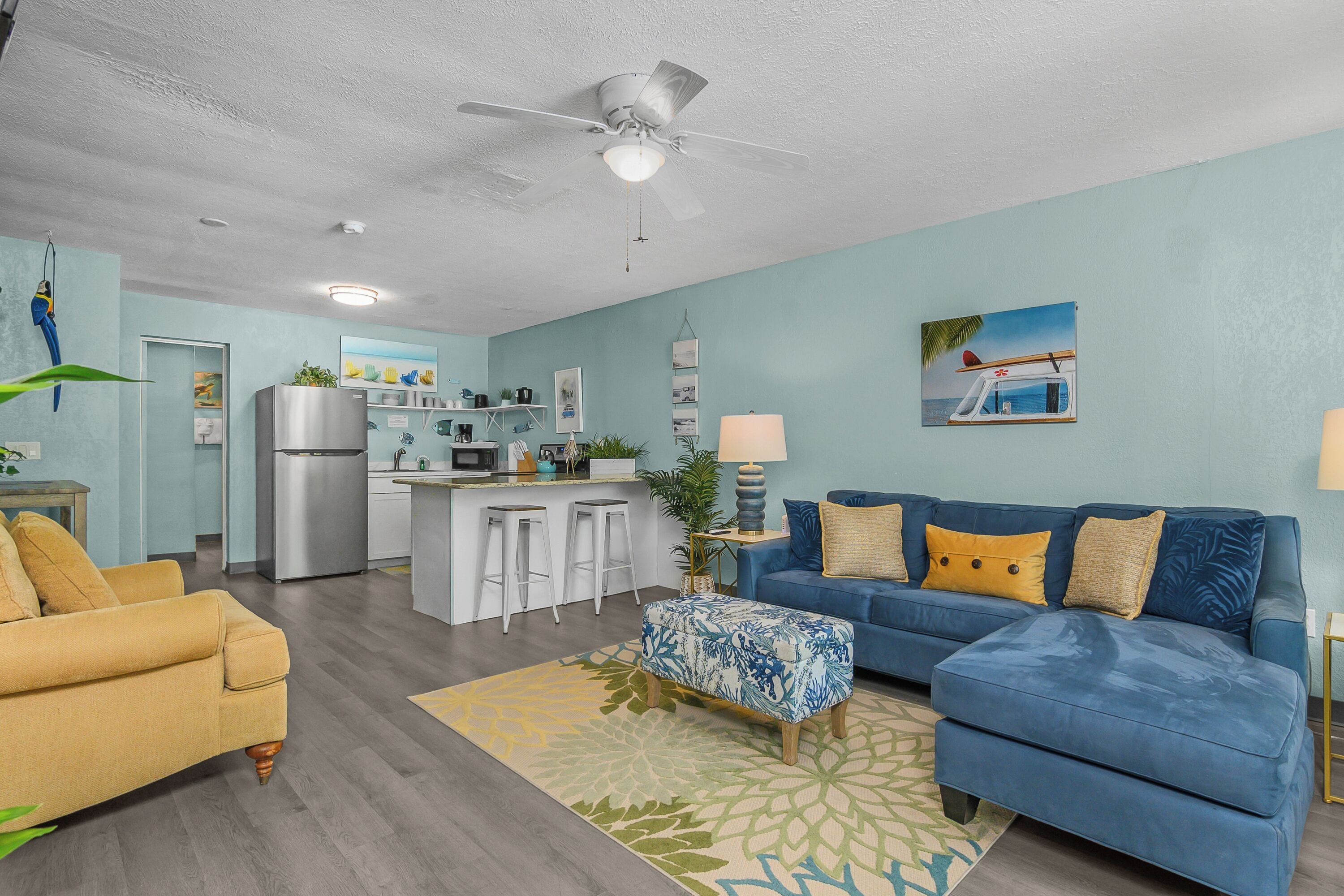 Una sala de estar con un sofá azul, un sillón amarillo y una zona de cocina con refrigerador y taburetes de bar.