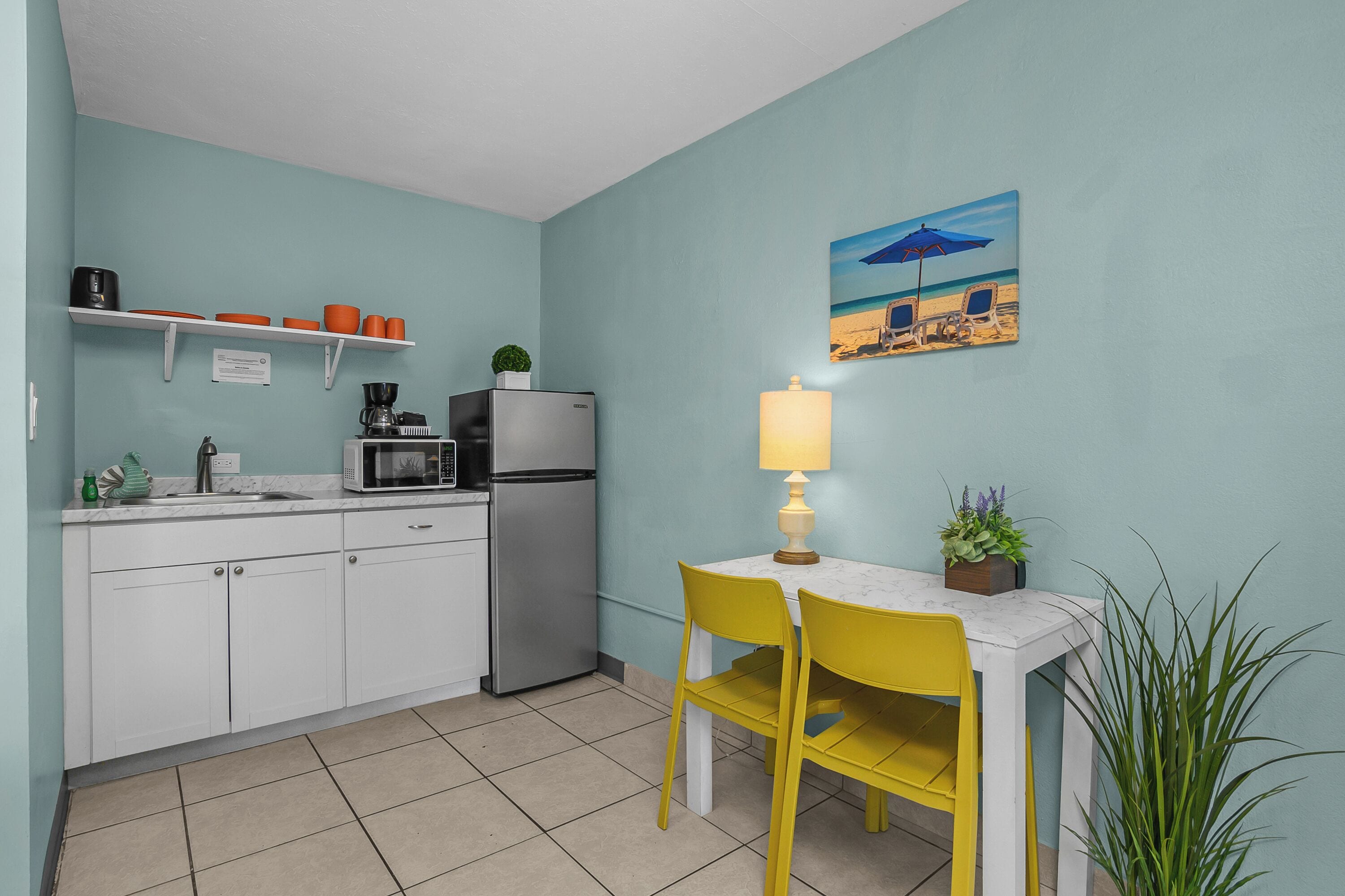 A kitchen with white cabinets, a refrigerator, a microwave, and a sink. There is a table with a yellow chair and a lamp. A painting of a beach scene hangs on the wall.