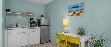 A kitchen with white cabinets, a refrigerator, a microwave, and a sink. There is a table with a yellow chair and a lamp. A painting of a beach scene hangs on the wall.