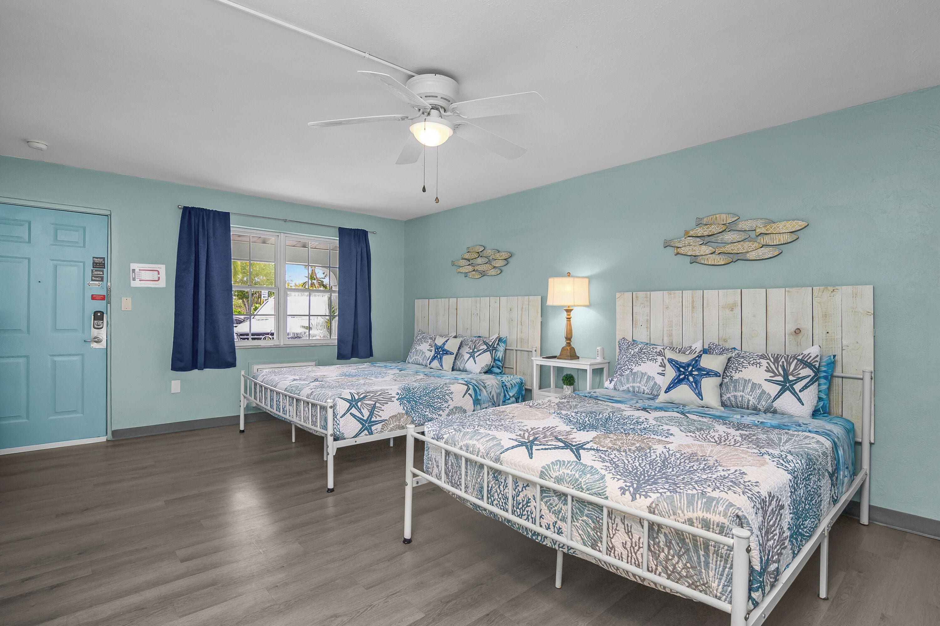 A room with two beds, a ceiling fan, a window with curtains, and a wall-mounted lamp.